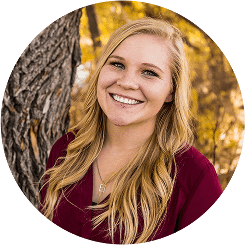 Smiling blonde woman in burgundy top, outdoor portrait with blurred autumn foliage background.