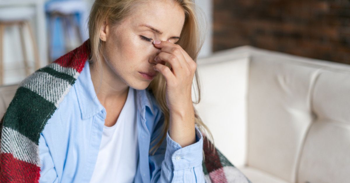 Woman with hand to face showing discomfort, wrapped in blanket on couch at home.