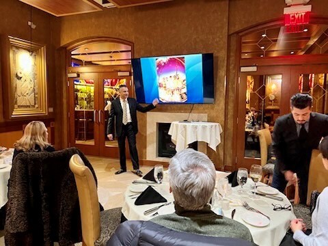 Formal event in elegant wood-paneled room with attendees dining and watching presentation on screen.