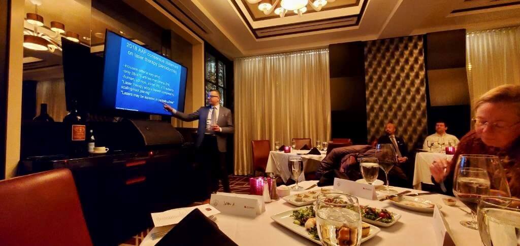 Dental professionals presenting at conference in elegant banquet hall with table settings and screen display.