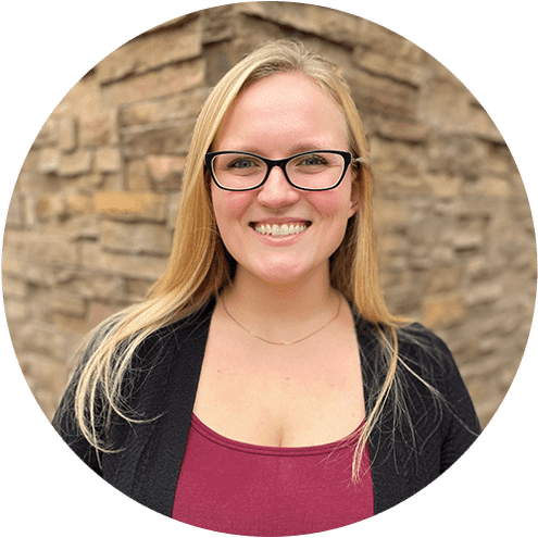 Professional headshot of smiling woman with glasses wearing maroon top against brick wall background.