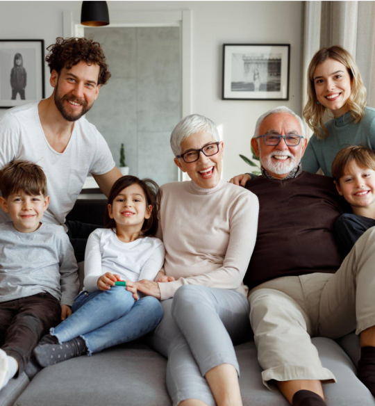 Multi-generational family sitting together on couch in living room, smiling at camera.