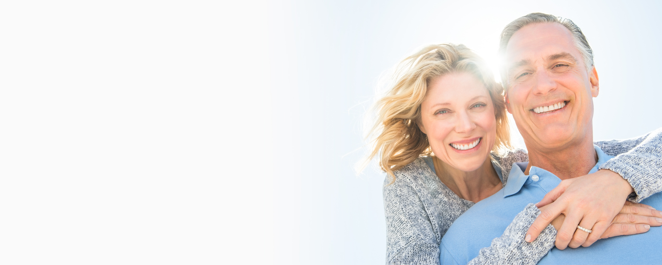 Middle-aged couple smiling together outdoors, embracing in casual clothing against light background.