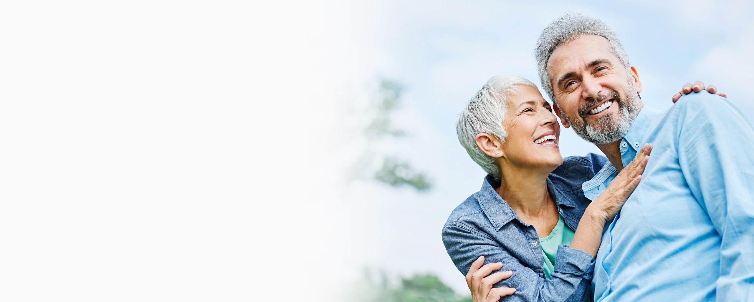 Older man and woman smiling outdoors, wearing casual clothing in bright daylight setting.
