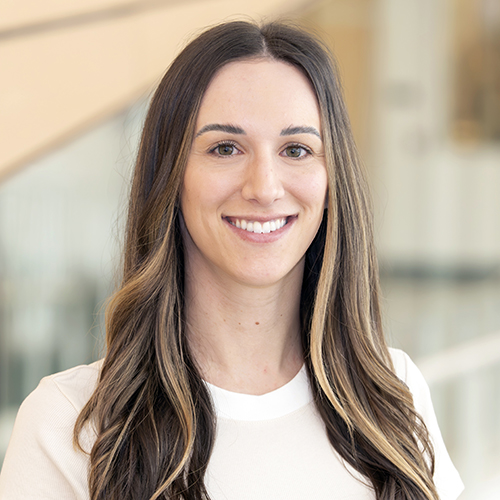 Professional headshot of smiling woman with long dark hair in white top, indoor office setting.
