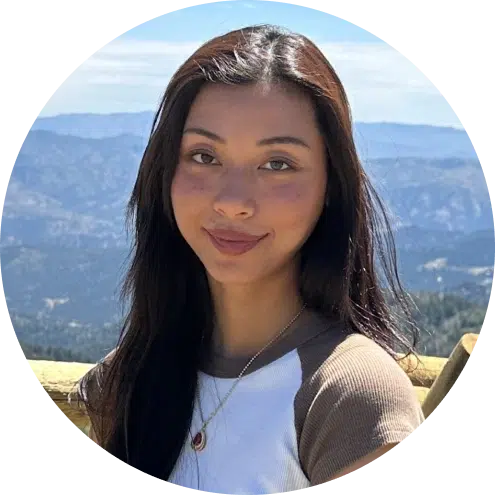 Woman smiling outdoors with mountain landscape background, wearing casual white shirt.